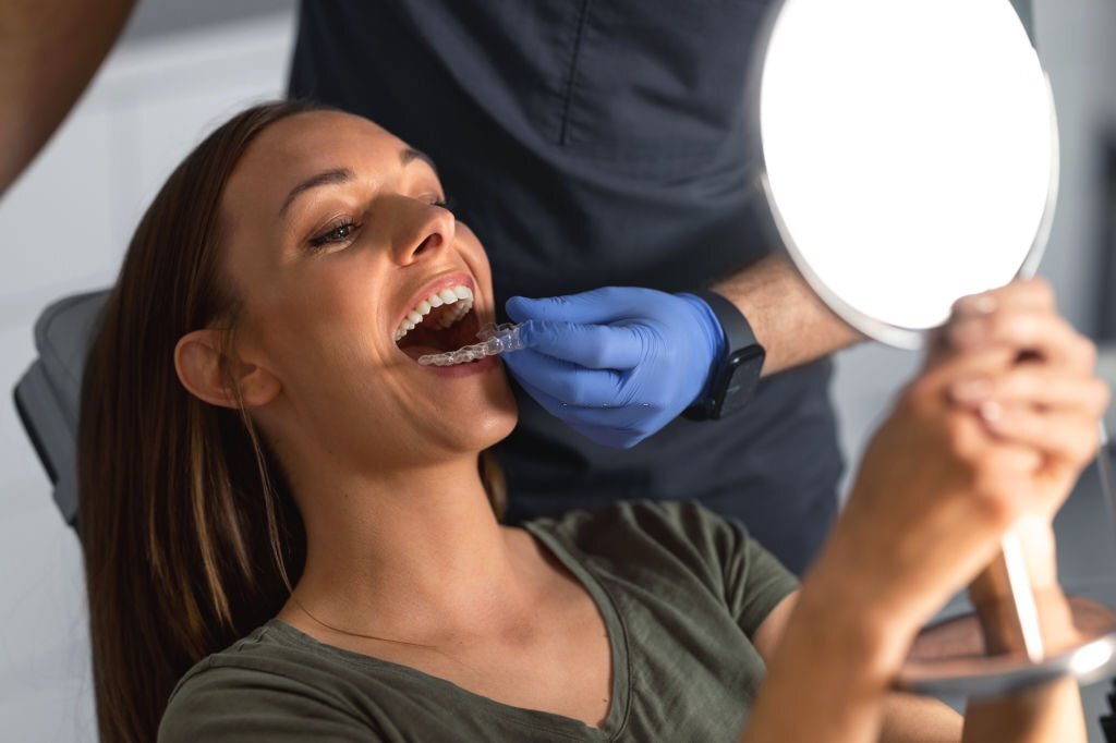 Close-up shot of a young woman looking at the hand mirror, checking her results after teeth whitening in the dentist's office while an unrecognizable dentist is removing the silicon tray for teeth whitening from her mouth.